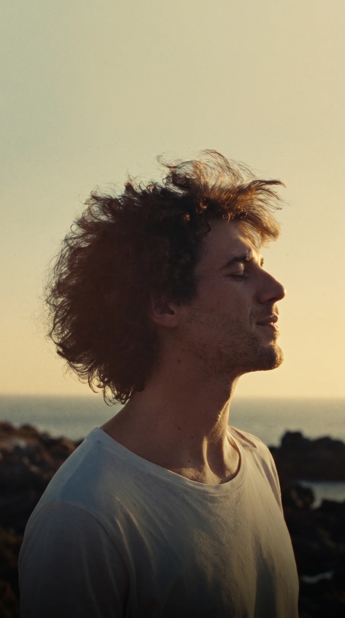 A man with a full head of hair, eyes closed, facing the evening sun by the sea